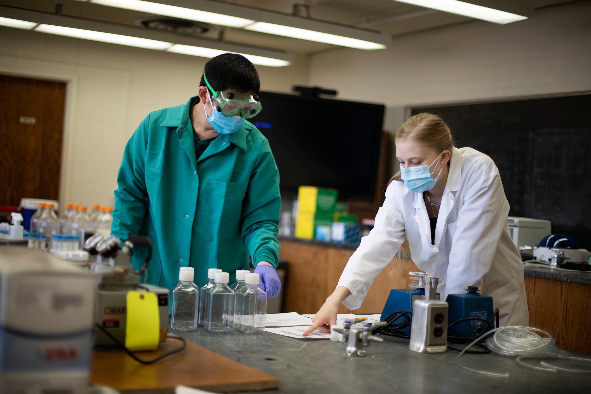 Professor and student in protective gear analyze data on paper beside empty sample tubes